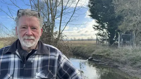 Greg Knowlson faces the camera with a small brook in the background. A sewage pipe is visible to his left, discharging clean water into the brook. A smaller pipe is located under the water which releases untreated water. Greg is wearing a large checked padded jacket, has a grey beard and grey hair with glasses on his head. 