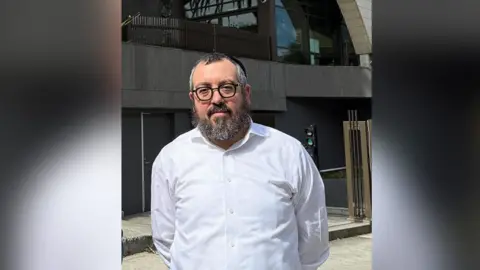 Social media Rabbi Eli Schlanger looks at the camera wearing a white shirt. He has grey hair, a beard and is wearing black-rimmed glasses. 