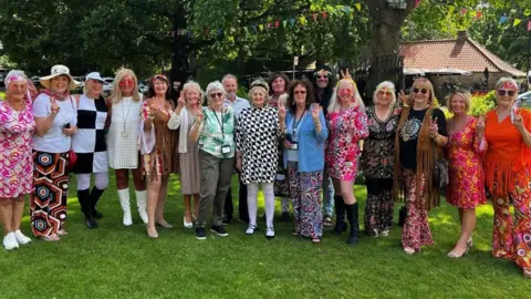 Washington in Bloom The Washington Bloom Group on the village green dressed in colourful 1960s costumes. There are trees and bunting in the background. They are smiling and many of them are doing a peace gesture. 