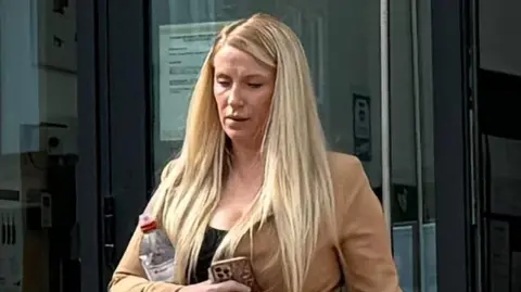 BBC A woman with long blonde hair tied up in a ponytail walks out of court looking down. She wears a black top with a beige jacket. She has a water bottle tucked under her arm and is holding an iPhone.