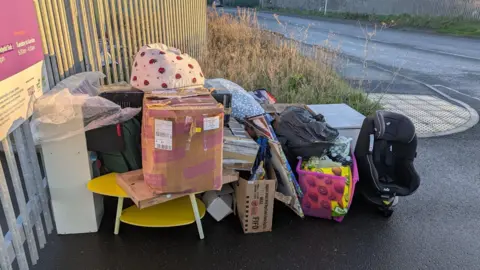 Enfys Foundation Piles of bags and boxes of donations outside the gate of the charity store including a baby car seat and yellow coffee table. 