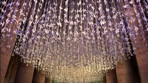 Thousands of paper doves hang from the ceiling of an abbey with purple lighting shining over them.