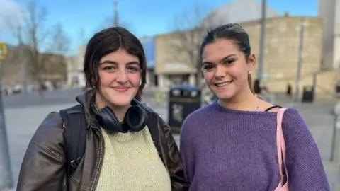 Olivia Gallosi is on the left and has brown hair, a yellow jumper and a brown jacket. She has headphones around her neck and a rucksack on her back. Roni Sharp is on the right and is wearing a purple jumper and has a pink bag on her shoulder. There are buildings in the background.