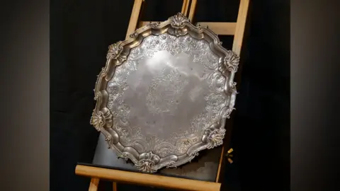 A large circular silver salver is rested on an upright wooden frame. It has a decorative finish.