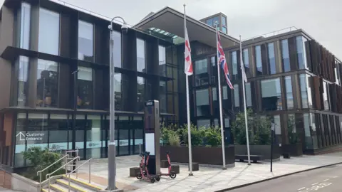 West Northamptonshire Council headquarters. It is a two-storey glass-fronted building with three flagpoles at the front.