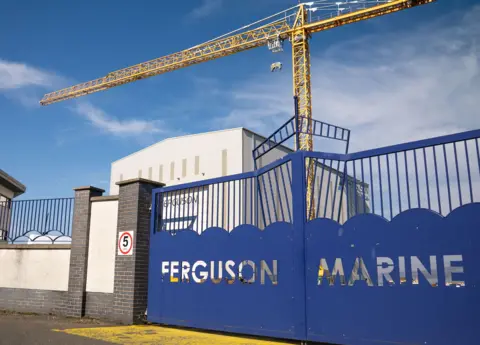 A blue gate with the words Ferguson Marine cut out. A large module hall and yellow crane can bee seen in the background