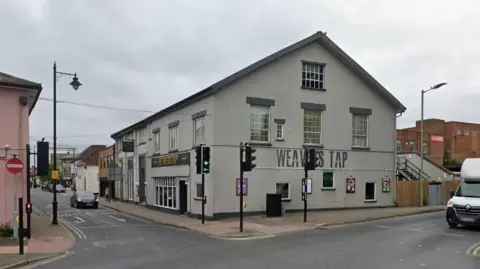 Google The Weavers Tap pub - a three-storey building on the corner of a town centre crossroads. It has a light grey painted exterior with dark grey lintels about the first and second floor windows. 