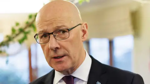 John Swinney, a bald man with glasses wearing a darks suit and white shirt, looks towards the camera in a close-up shot 