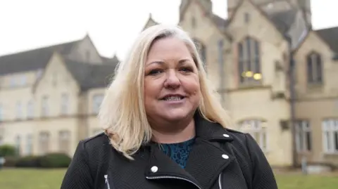 John Fairhall/BBC Joanna Hudson is standing in front of a large Victorian brick building, which is blurred in this image. She has shoulder-length blond hair and is wearing a blue top and a black jacket