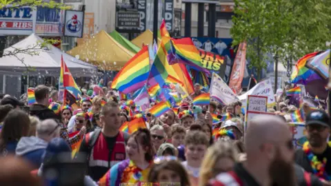 A crowd of people waving rainbow flags with a few trees and large buildings behind them