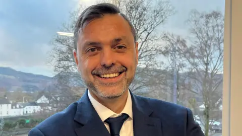 Martin Birch smiling at the camera. He has short salt and pepper hair and dark eyes. He is wearing a blue suit and tie and a white shirt.