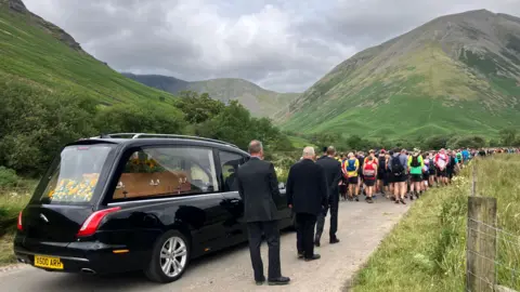 Hundreds of runners wearing club colours lead Joss Naylor's funeral cortege in the Lake District with fells in the background