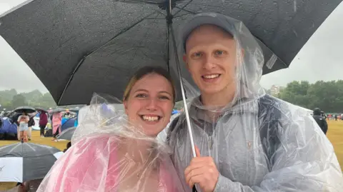 Emily Coady-Stemp/BBC A man and woman, wearing transparent waterproofs, stand under an umbrella