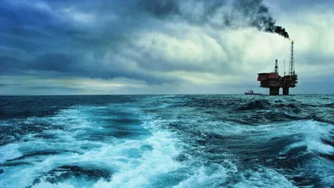 Getty Images An oil rig in the North Sea, on the far right of the shot with choppy waves in the foreground. Black smoke is billowing from a chimney on the rig, with a boat behind it.