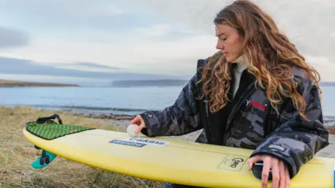 Pete Hill A person is sitting on grassy dunes by the sea, holding a yellow surfboard and applying wax to its surface. The background shows calm water and a distant coastline under a cloudy sky, with a dark jacket and a white jumper visible on the person, and surfboard branding stickers on the board.