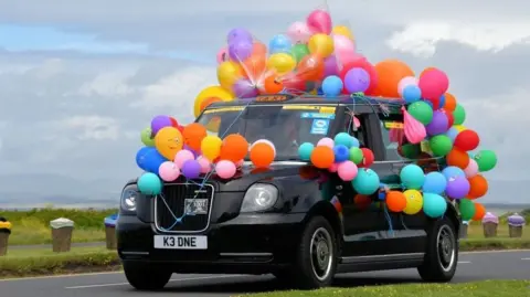 A black taxi covered in bright balloons as part of a charity event.