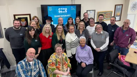 Shrewsbury Town Council A group of 20 people smiling at a camera inside a council room with a big screen behind them with the word "Shrewsbury" written in white on a blue background.