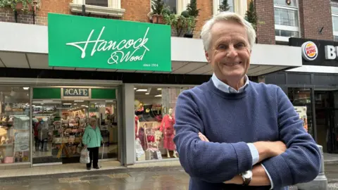 A grey haired smiling Christopher Hancock stands, arms folded in a blue jumper over a white shirt, in front of the green-signed Hancock and Wood departments store 