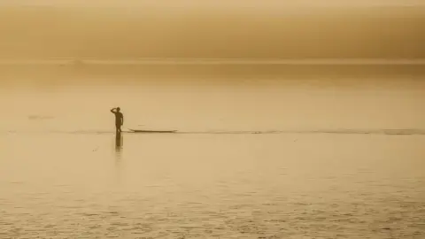 A surfer salutes on the still water at dusk as he stands next to his surfboard. The sky and the river appear orange due to the haze.