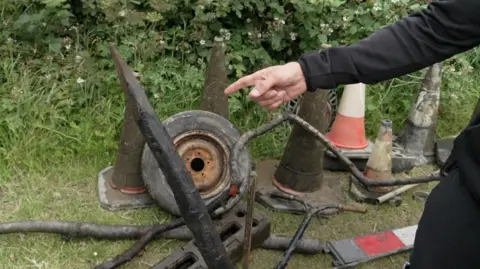 A hand points towards a sharp wooden stake which divers have pulled out of the water. Alongside it on the bank is a collection of debris. The items include a rusted wheel, traffic cones, and a scooter's handlebars. 