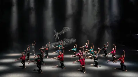 Johan Presson a companhia de dança usando calças pretas e camisas coloridas em um palco preto iluminado.