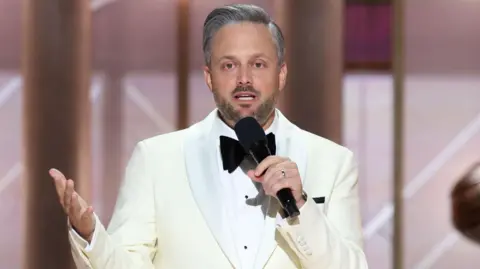 Getty Images Nate Bargatze pictured wearing a white tuxedo and holding a microphone, during the 82nd Annual Golden Globes held at The Beverly Hilton on January 05, 2025 in Beverly Hills, California