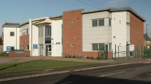 BBC/Ed Young A modern, two-story building with a combination of red brick and white panel exterior walls. The structure has a contemporary design featuring clean lines and a slightly curved roof section on the left side. Large windows are visible on both floors, allowing natural light into the building. The main entrance is located at the centre, marked by a glass door and flanked by blue signage. There is a paved pathway leading to the entrance, bordered by a small landscaped area with greenery. Surrounding the building is a metal fence, and the foreground includes a road with a pedestrian crossing. In the background, there are residential houses and leafless trees.