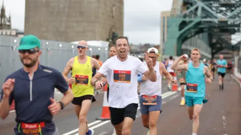 North News Half a dozen runners crossing the Tyne Bridge. The men are wearing running gear, with white, turquoise, dark blue and yellow vests, and turquoise, black and blue shorts. The runner in the middle is holding up his left hand in a victory sign.