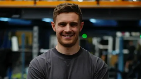 Rowmads A man wearing a grey T-shirt while standing in a gym. He has a beard and is smiling at the camera.
