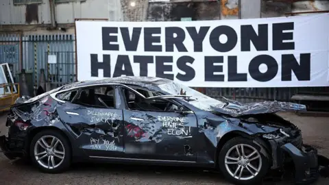 Getty Images A banner reading "everyone hates Elon" behind a smashed up Tesla