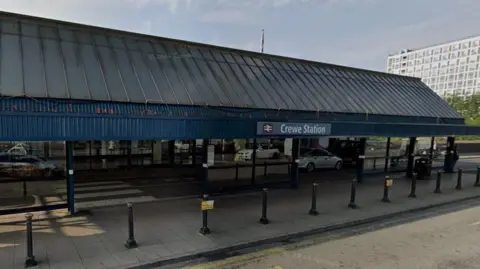 The outside of a railway station. It is a single storey building with a blue slanted roof and a sign that says Crewe Station. There are bollards lining the pavement in front of the building