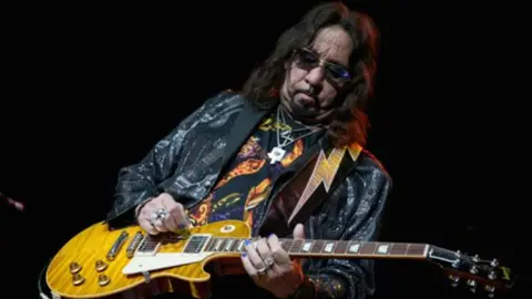 Getty KISS guitarist Ace Frehley plays an electric guitar with a lightning-strike guitar strap. 