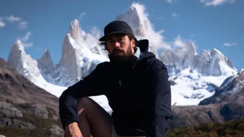 Charlie Probets Mike Humphreys sat on a mountain wearing a black cap, a black coat and shorts. A large white mountain is behind him. 
