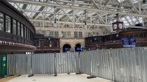 The main waiting area of Glasgow Central by the Gordon Street entrance. The waiting area is obscured by metal sheeting and there is a glass roof held up by white barring. The place is empty