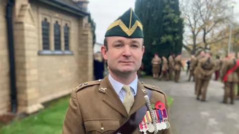 Lieutenant Colonel Gerald Kearse, pictured in uniform outside the church in Tidworth. His is wearing a light brown suit with multiple medals and a poppy attached, and is looking slightly away from the camera. 