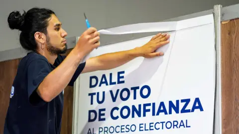 An election worker with dark hair tied in the back, a beard and a large earring is seen in profile as he sticks a poster at an election centre