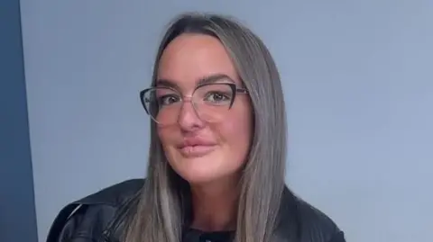 Toni Garrigos poses for a picture against a grey wall. She has long fair hair and wears glasses plus a dark jacket.