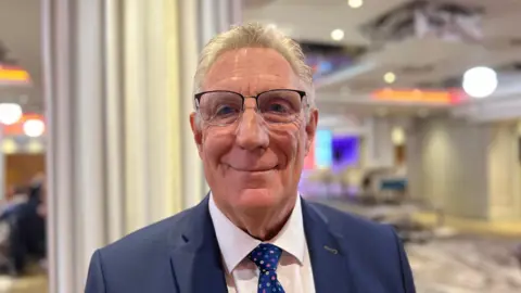 BBC Malcolm Lewis is wearing a blue blazer and a blue tie with flowers. He has glasses on. He is pictured in a conference room. 