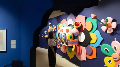 Benedict Johnson A woman looking at a 3D piece of art featuring brightly coloured masks and flowers