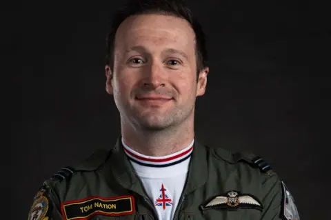 RAF Cosford Flt Lt Tom Nation wears a green RAF flight suit with his name badge on his right and the RAF insignia on the left of his jacket. He has a white vest on with red, white and blue collar. 