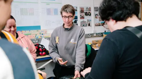 Caroline Henn teaches one of her DIY courses. She is looking down at a piece of wood. Other students are standing around her looking down as well. She has a pencil in her hand