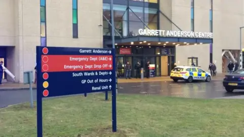 BBC News The entrance to the emergency department at Ipswich Hospital. A sign saying "Garrett Anderson Centre" is over the main door. There are people milling about and a police car. In the foreground there is a hospital sign with directions to wards and departments. 