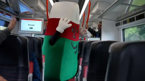 The red, green and white triangle character aboard the new Transport for Wales train. He is standing in the aisle with his hands in the air. 