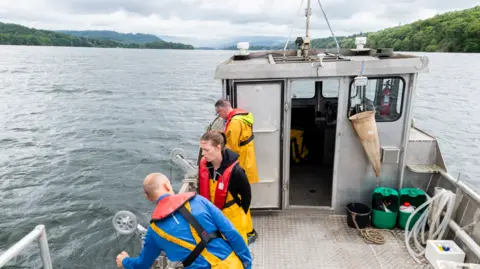 Paul Adams Photography A group of scientists wearing yellow fishing overalls and red life jackets stand on a monitoring motor boat. The boat has silver corrugated floor. The three scientists stand to the port-side of the boat and are fetching data from Windermere. 