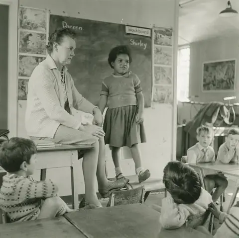 Estate of John Blakemore provided by Coventry University An classroom with four students sat as a desk looking at a teacher, who is sat on a desk with another student standing on a chair next to her.