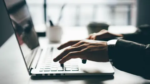 Computer user's hands on the keyboard of a laptop, which is sitting on a desk near a window.