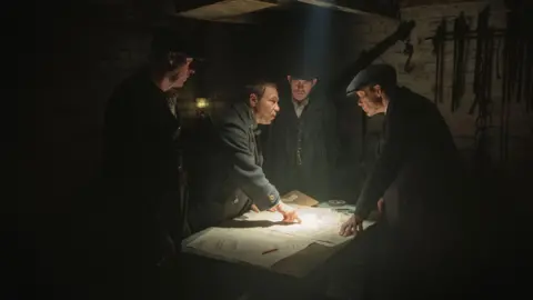 Netflix Several men in old fashioned suits are standing around a table in a basement and appear to be looking at a map which is lit up by an overhead light. One of the men on the left is pointing at the map.