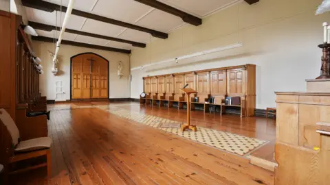 Hunters Estate Agents Inside one of the rooms. It has cream walls and brown wooden beams across a white ceiling. In the middle of the room is a stand with a microphone and what looks like an altar at the front with individual prayer stools down the side. At the back of the room are two large wooden doors with a cross on the top.