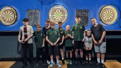 Members of Shooters Leominster Junior Darts Academy, wearing green club T-shifts, shorts and trainers, with darts boards behind them.