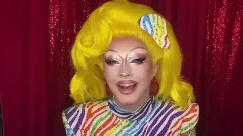 BBC Charra Tea wears a bright yellow wig and dramatic neutral make up. She is wearing a multi-coloured zebra striped body suit with shoulder pads.